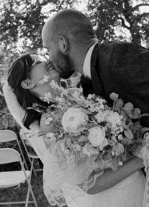 Black and white photo of a bride and groom sharing a kiss, bride holding a lush floral bouquet at an outdoor wedding.