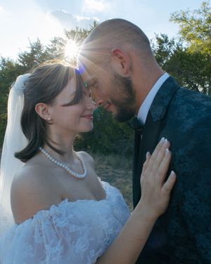 Bride and groom in romantic forehead-to-forehead pose outdoors, sun flare behind them, white dress and dark suit.