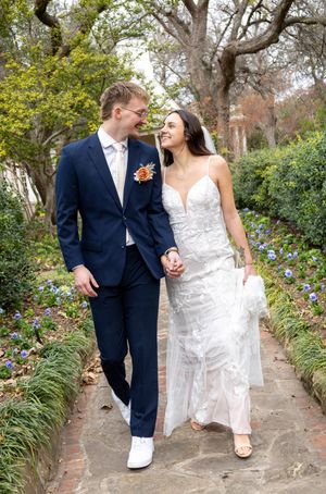 A bride and groom walk hand in hand through a garden path, smiling at each other on their wedding day.