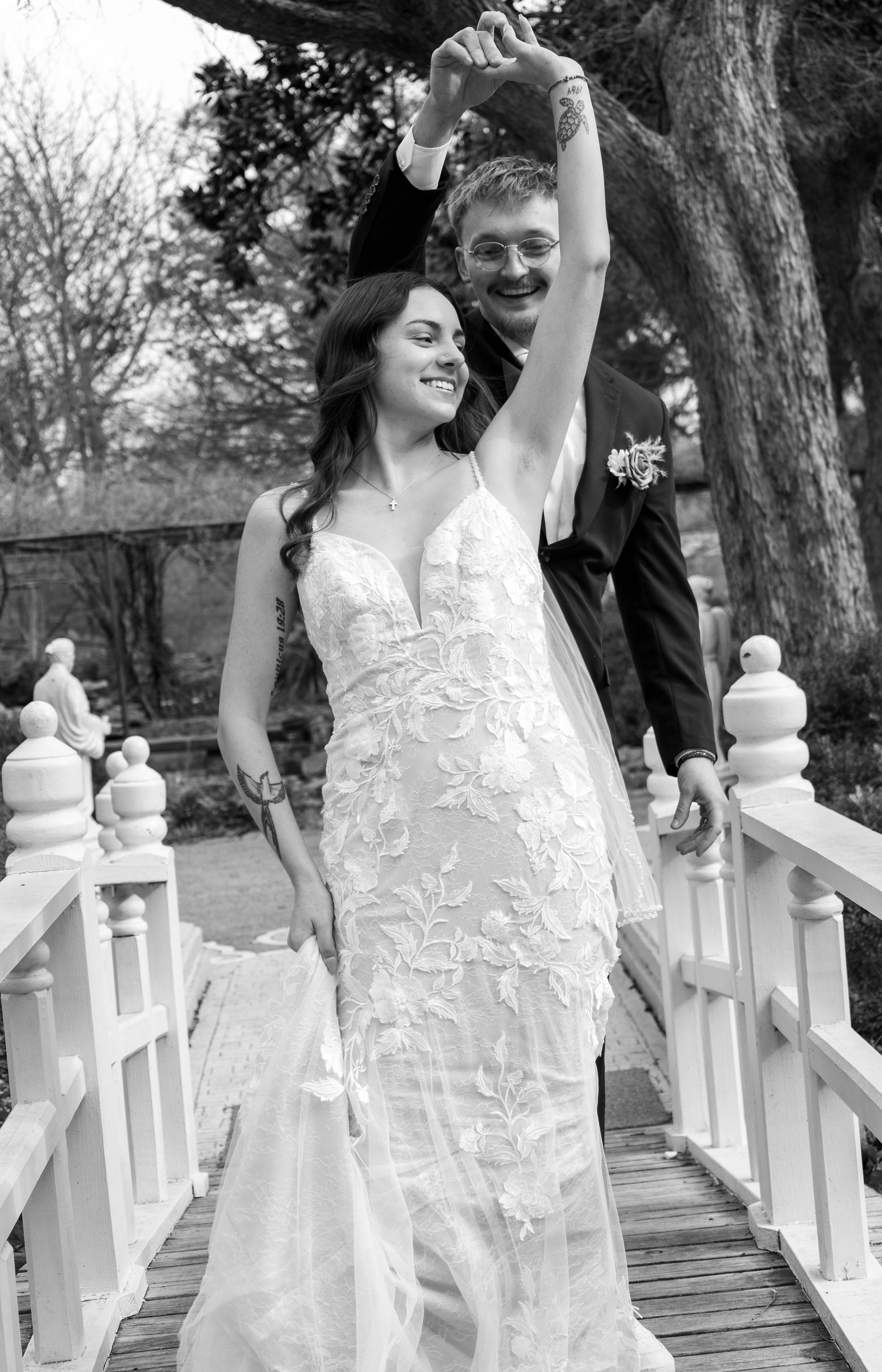 Black and white photo of a bride in lace gown and groom in tuxedo celebrating on a white bridge.