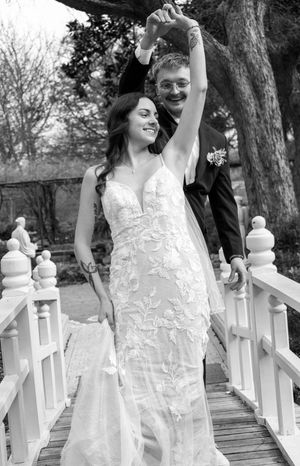 Black and white photo of a bride in lace gown and groom in tuxedo celebrating on a white bridge.