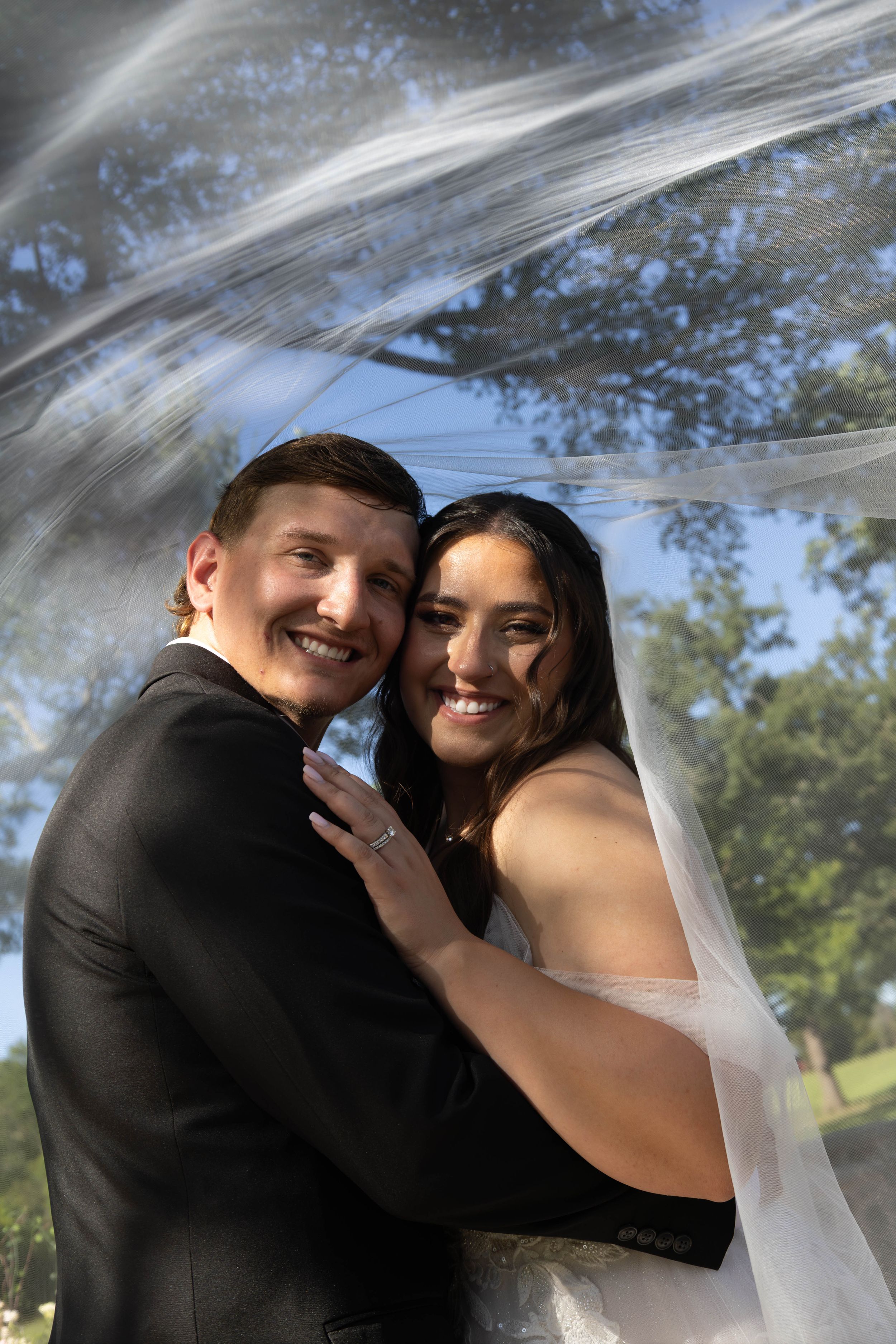 A smiling bride and groom embrace outdoors under a flowing wedding veil with blue sky and trees in the background.