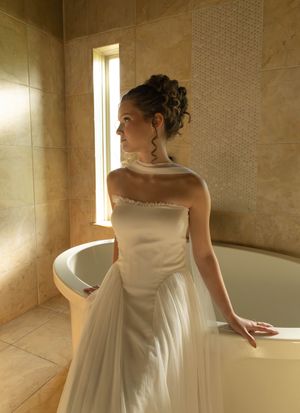 Elegant bride in white strapless wedding gown standing beside a luxury bathtub in a marble bathroom.