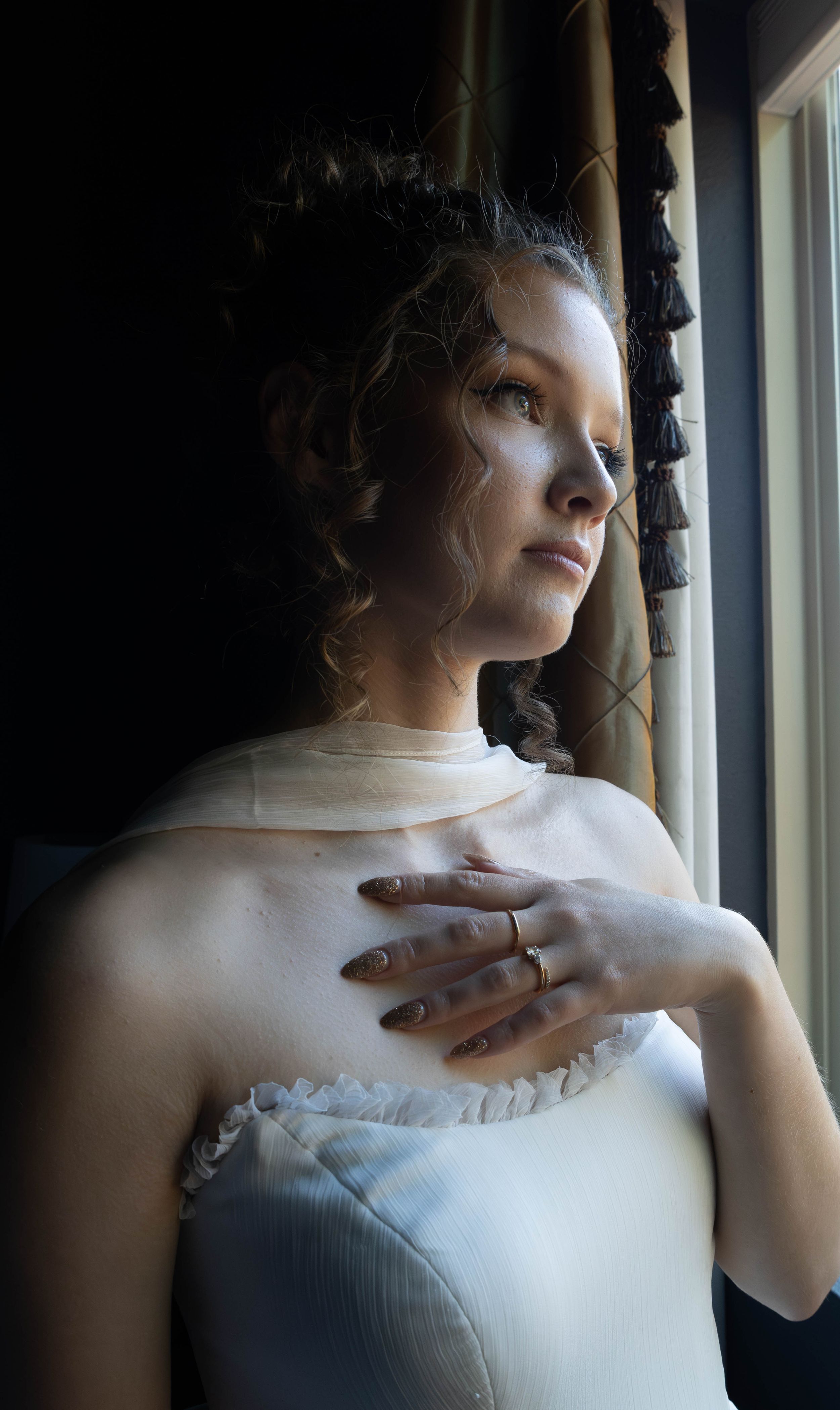 A woman in a white period dress gazes out a window, hand on chest, with elegant upswept curly hair.