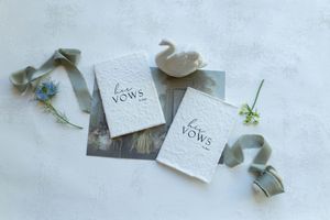 Two elegant white vow books with calligraphy lettering, styled with a ceramic swan, blue flowers, and sage ribbon.