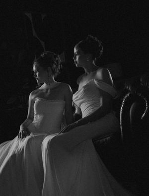 Two women in elegant white gowns sit together in dramatic black and white portrait photography.