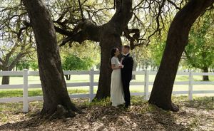 Couple in wedding attire standing romantically under large oak trees beside a white fence outdoors.
