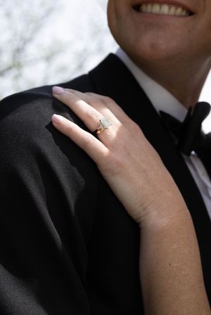 Close-up of a woman's hand with a gold diamond engagement ring resting on a man's black tuxedo shoulder.