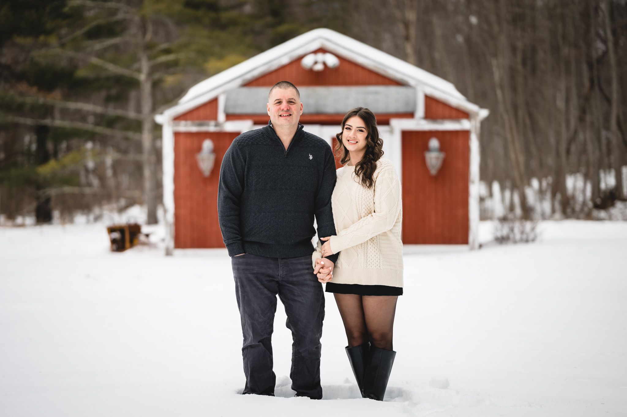Couple holding hands in snow before red barn in winter.