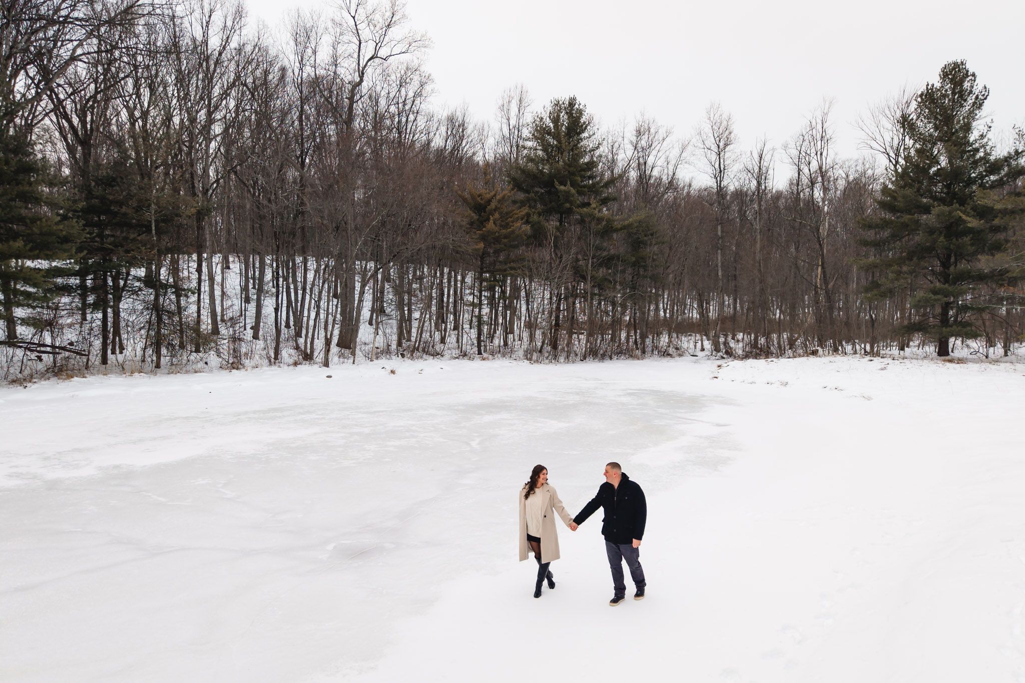 Couple holding hands walking across a snowy frozen lake surrounded by bare winter trees.