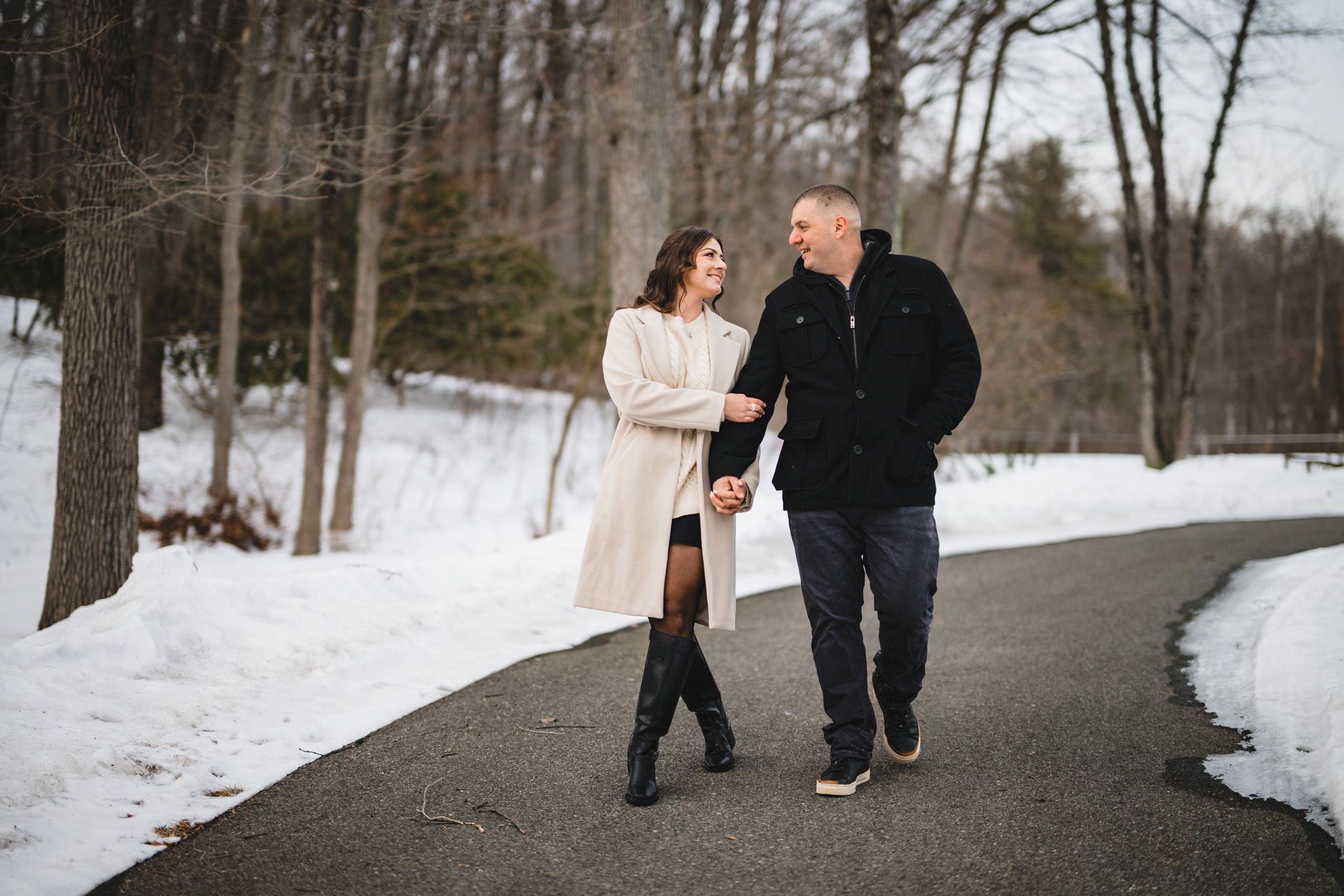 Couple holding hands and walking on a snowy park path in winter, woman in white coat and black boots.