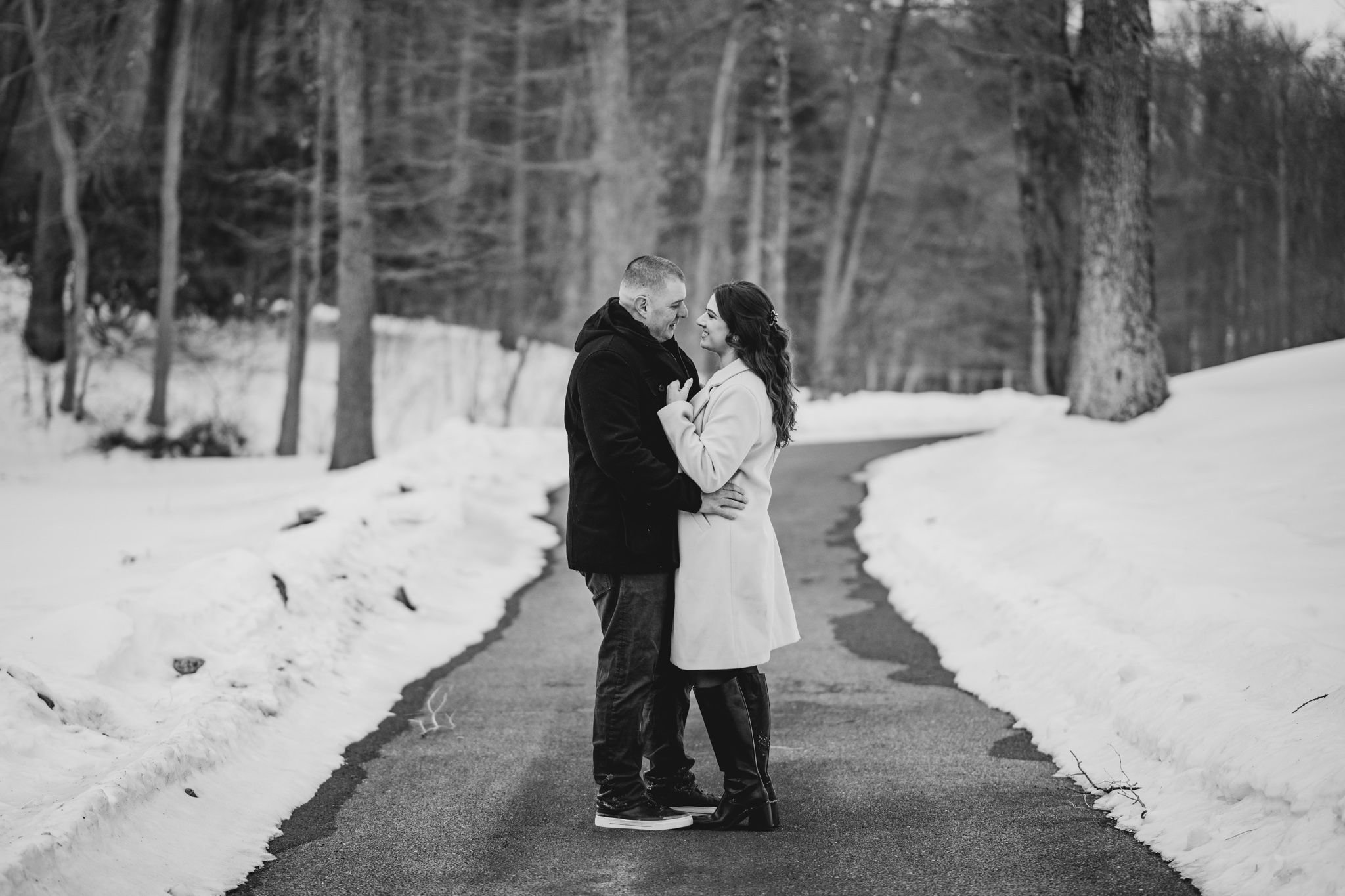 Black and white photo of a couple kissing on a snowy road surrounded by winter trees.