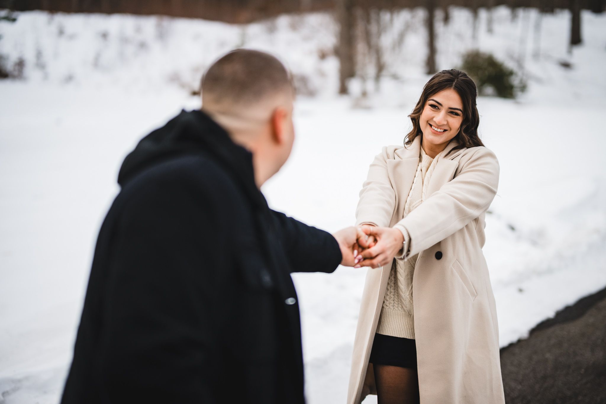 A smiling woman in a beige coat holds hands with a man outdoors in a snowy winter setting.