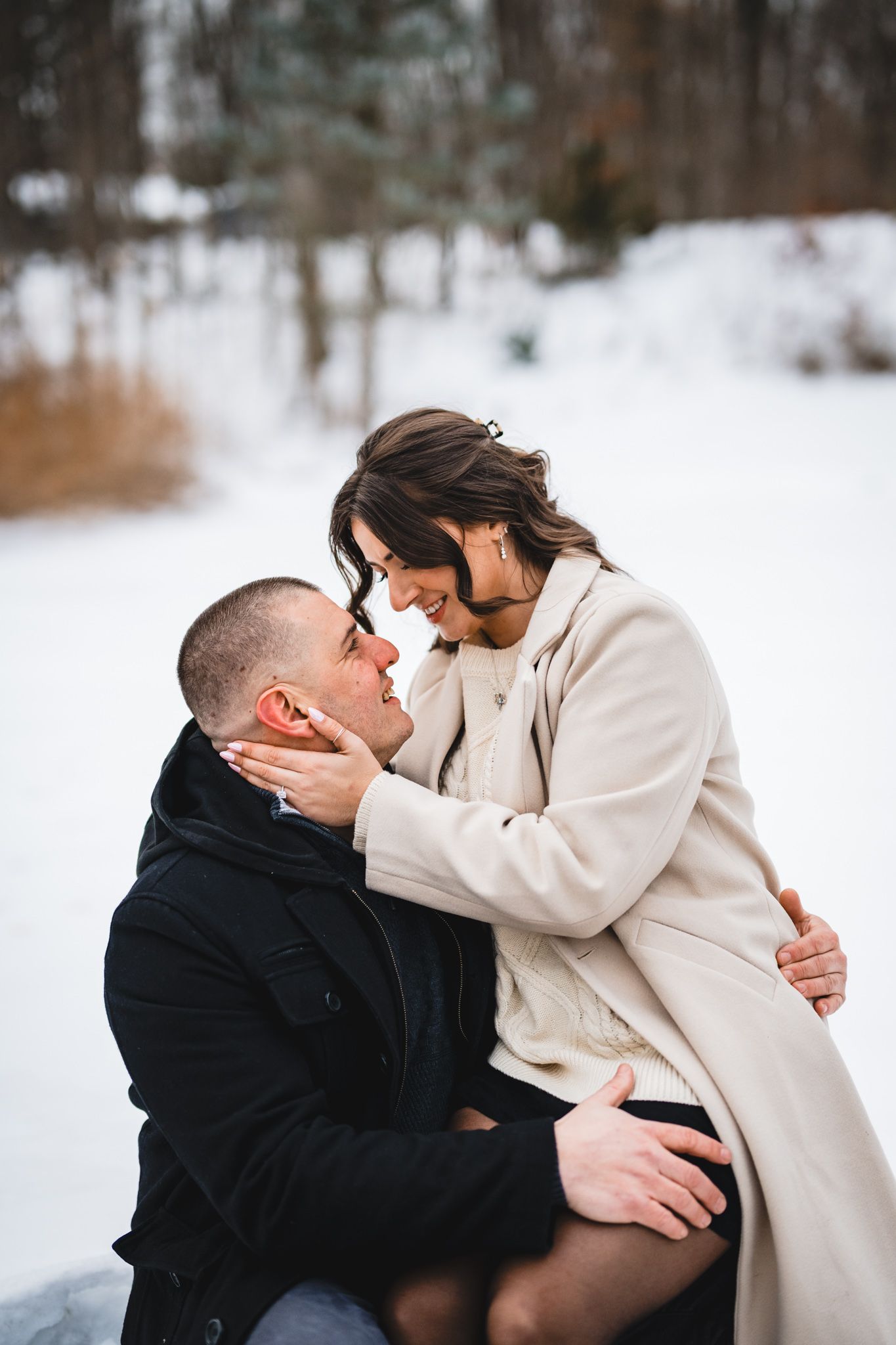Romantic couple sharing an intimate moment in a snowy winter forest, woman in beige coat leaning toward man in black jacket.
