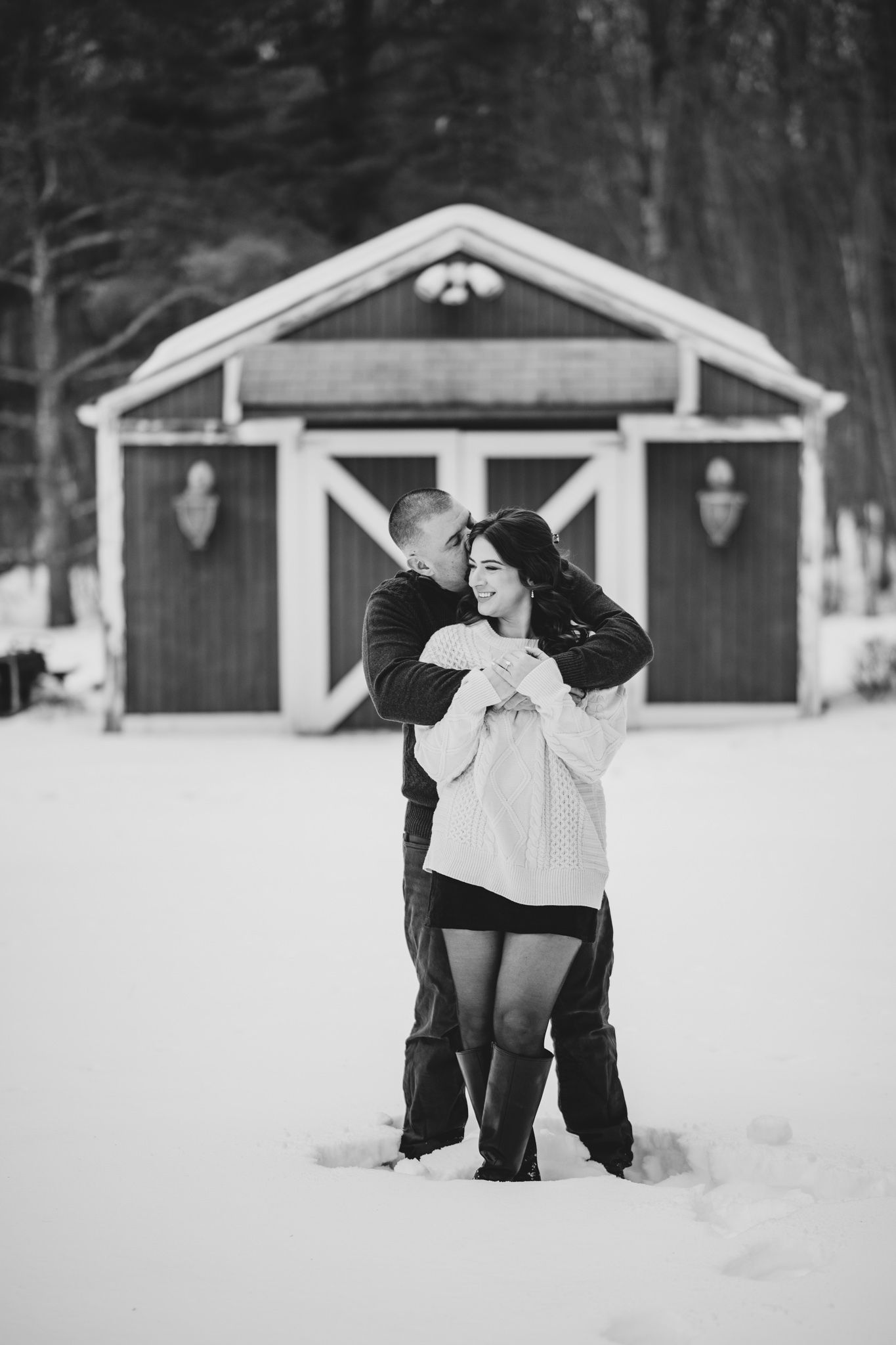 Couple embracing in snow before a rustic barn in black and white winter engagement photo.