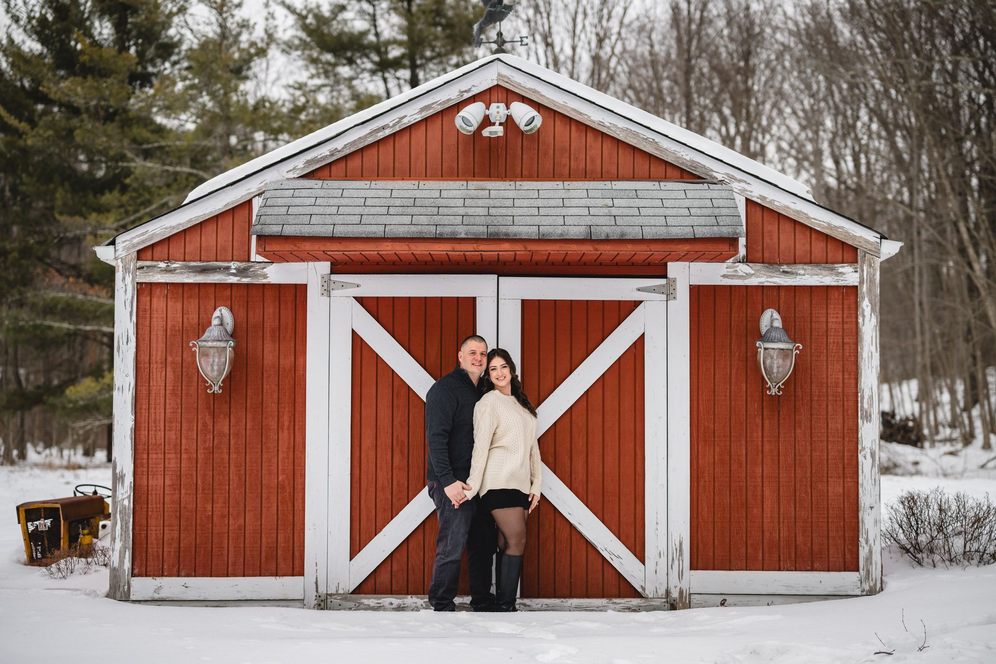 Couple embracing in front of red and white barn doors in snow.