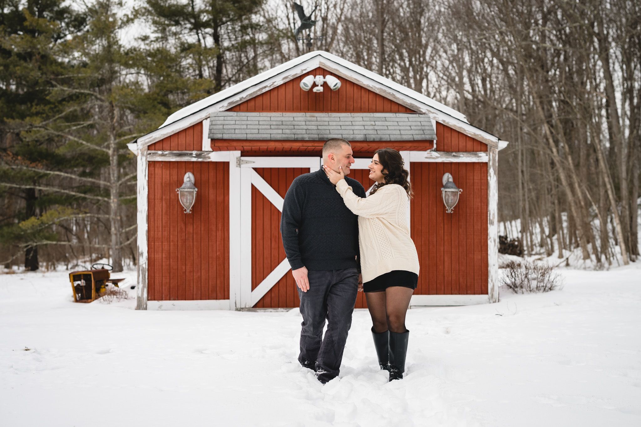 Couple embracing in snow before red barn in winter wonderland setting.