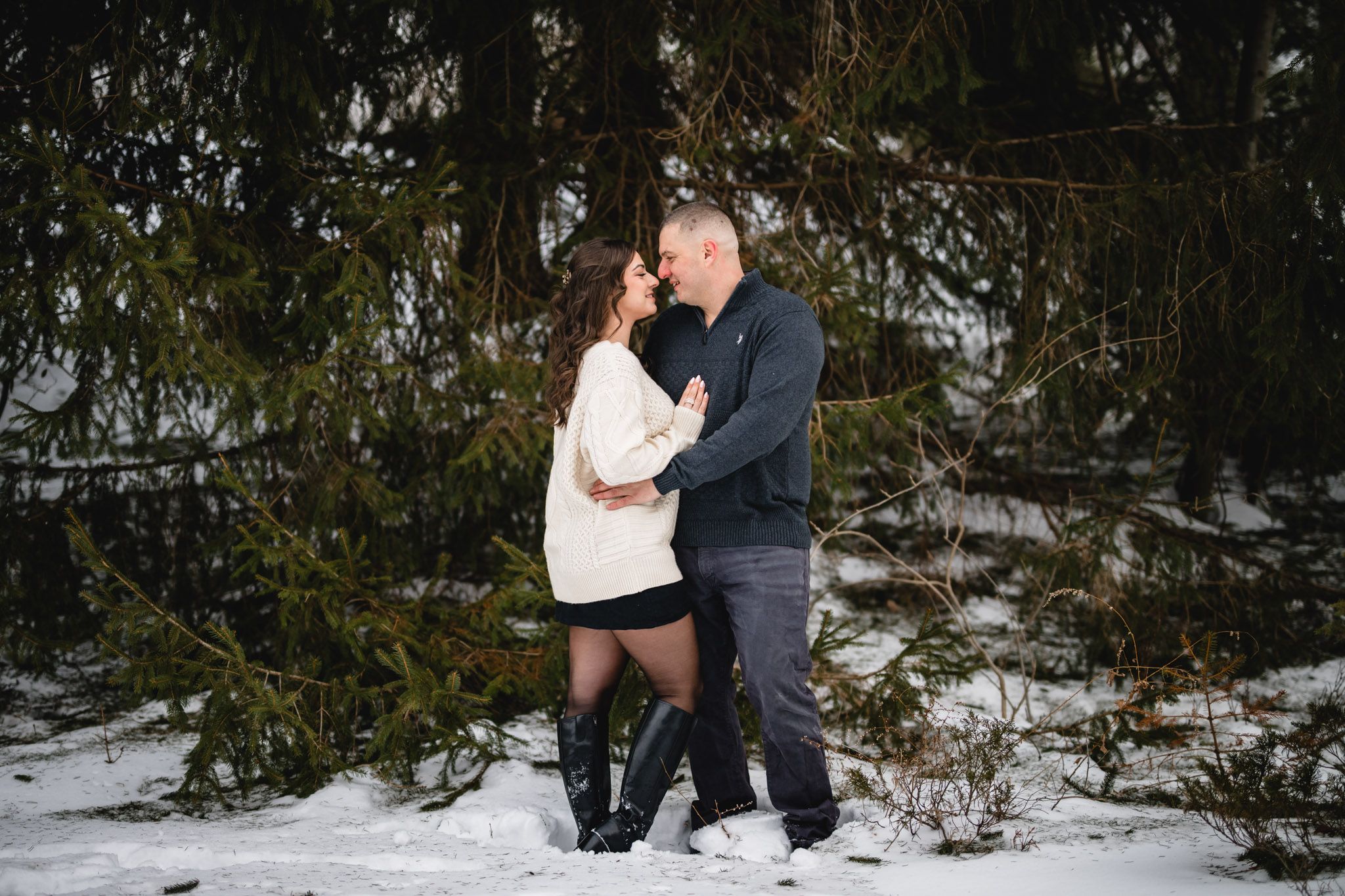 Couple embracing romantically in snowy winter forest with evergreen trees, wearing cozy sweaters during outdoor engagement photo session.