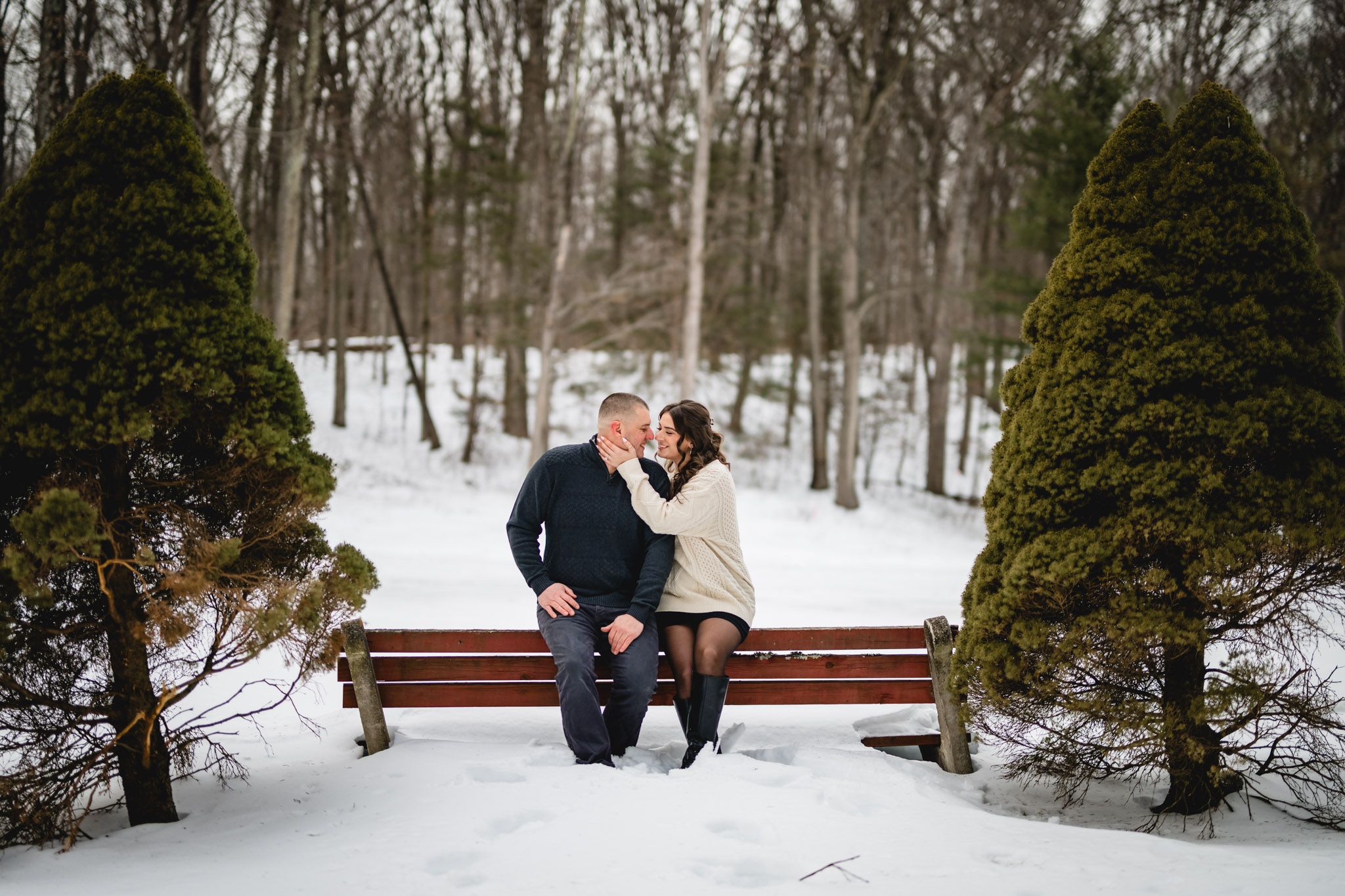 Couple sharing a romantic moment on a red bench in a snowy winter park surrounded by trees.