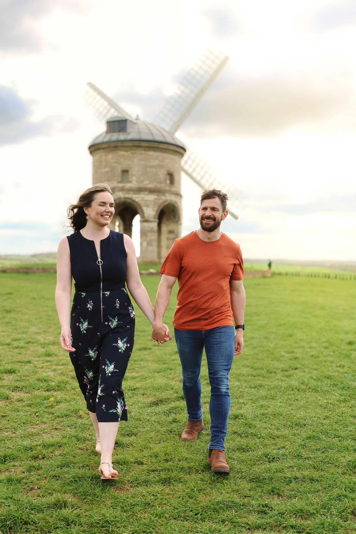 Couple embracing beside a historic stone windmill with green fields and cloudy sky in the background.