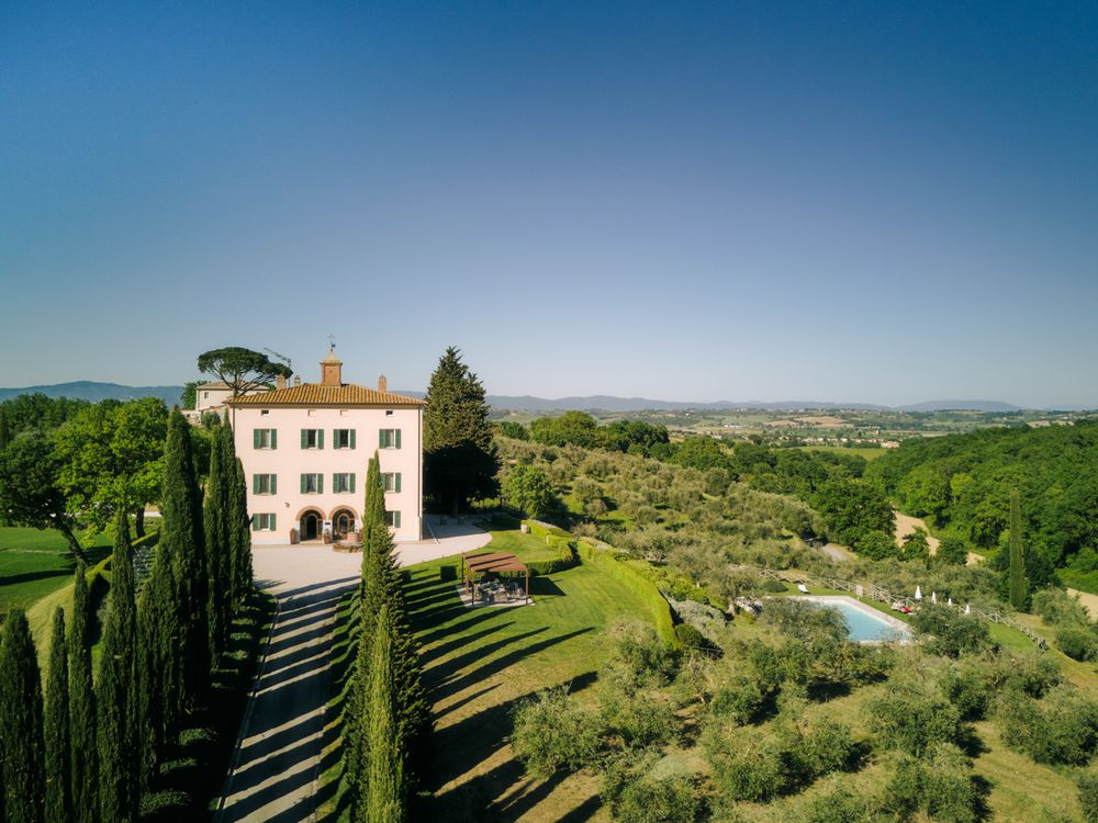 Aerial view of a historic Italian villa surrounded by cypress trees, lush gardens, and rolling Tuscan countryside under a clear blue sky.