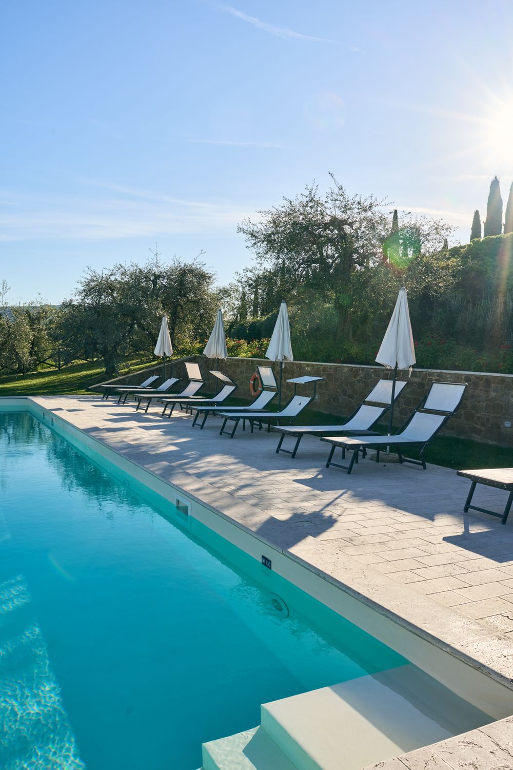 Luxury resort pool with white lounge chairs and closed umbrellas surrounded by lush greenery on a sunny day.