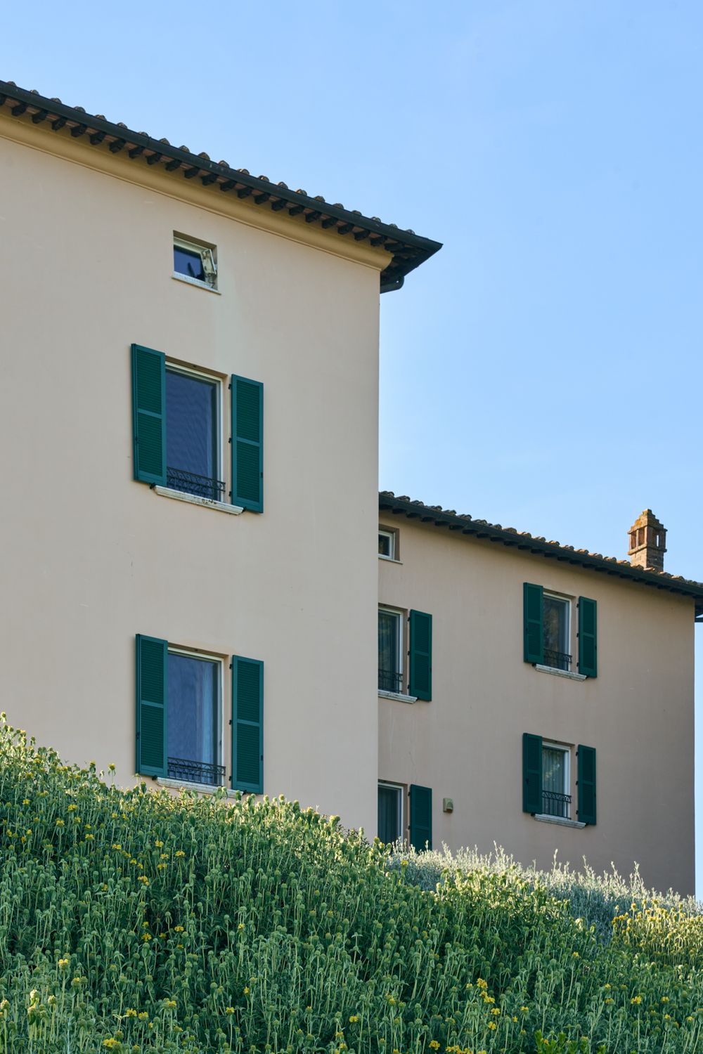 Tuscan-style building with green shutters and cream walls surrounded by lush greenery under a clear blue sky.