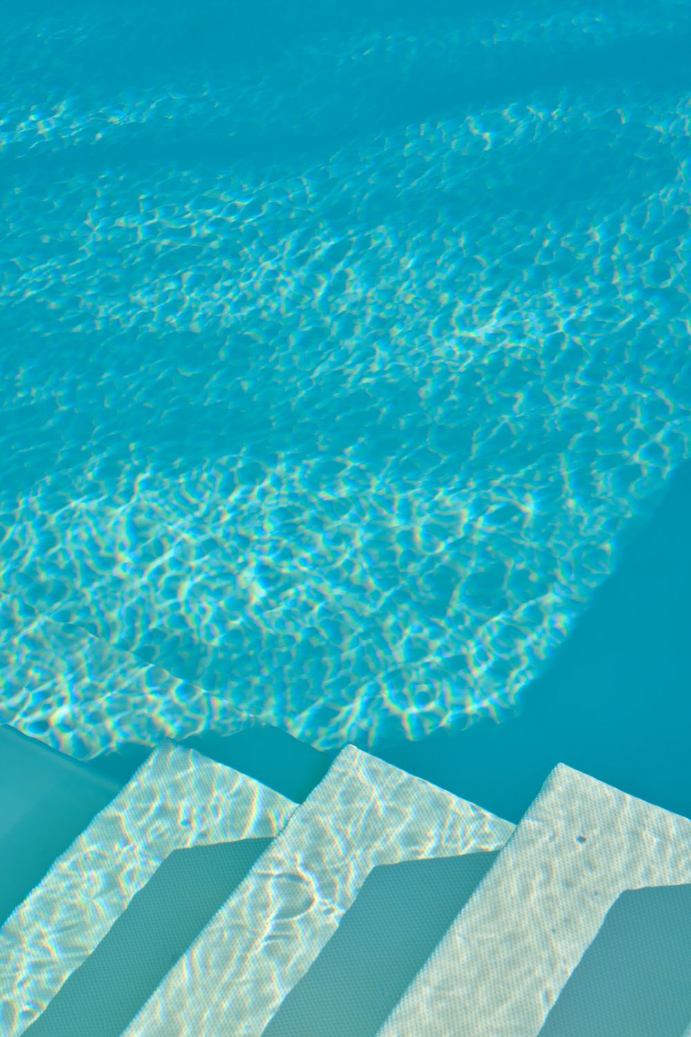 Crystal clear turquoise swimming pool with white steps leading into the shimmering water on a sunny day.