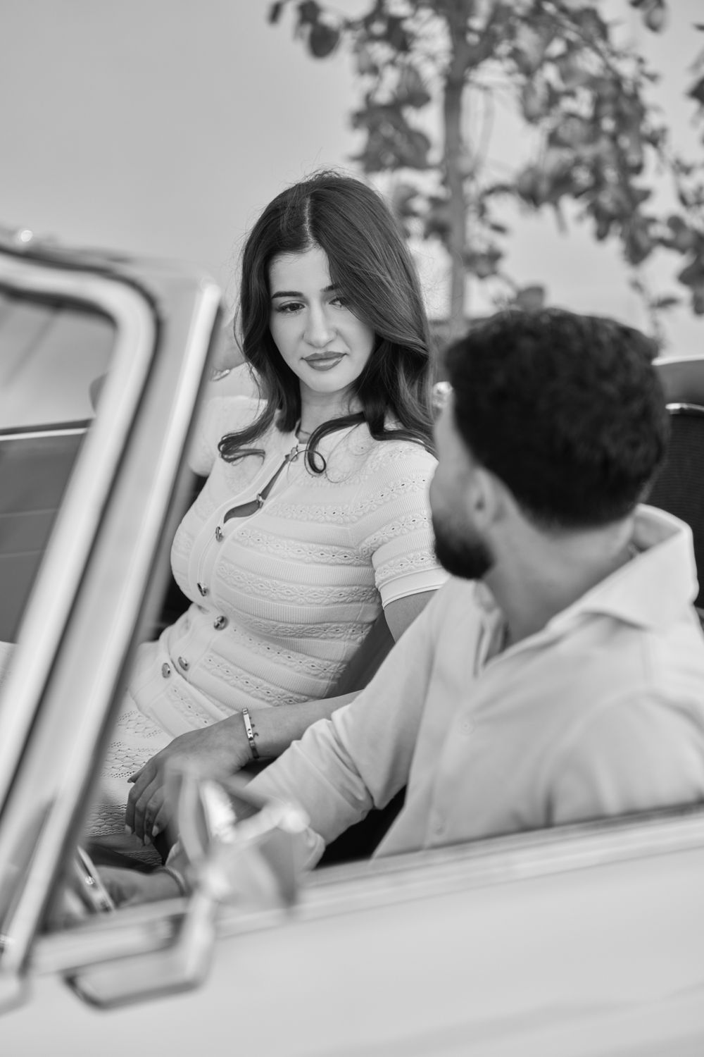 Black and white photo of a couple near a vintage car, woman in fitted dress smiling at bearded man.