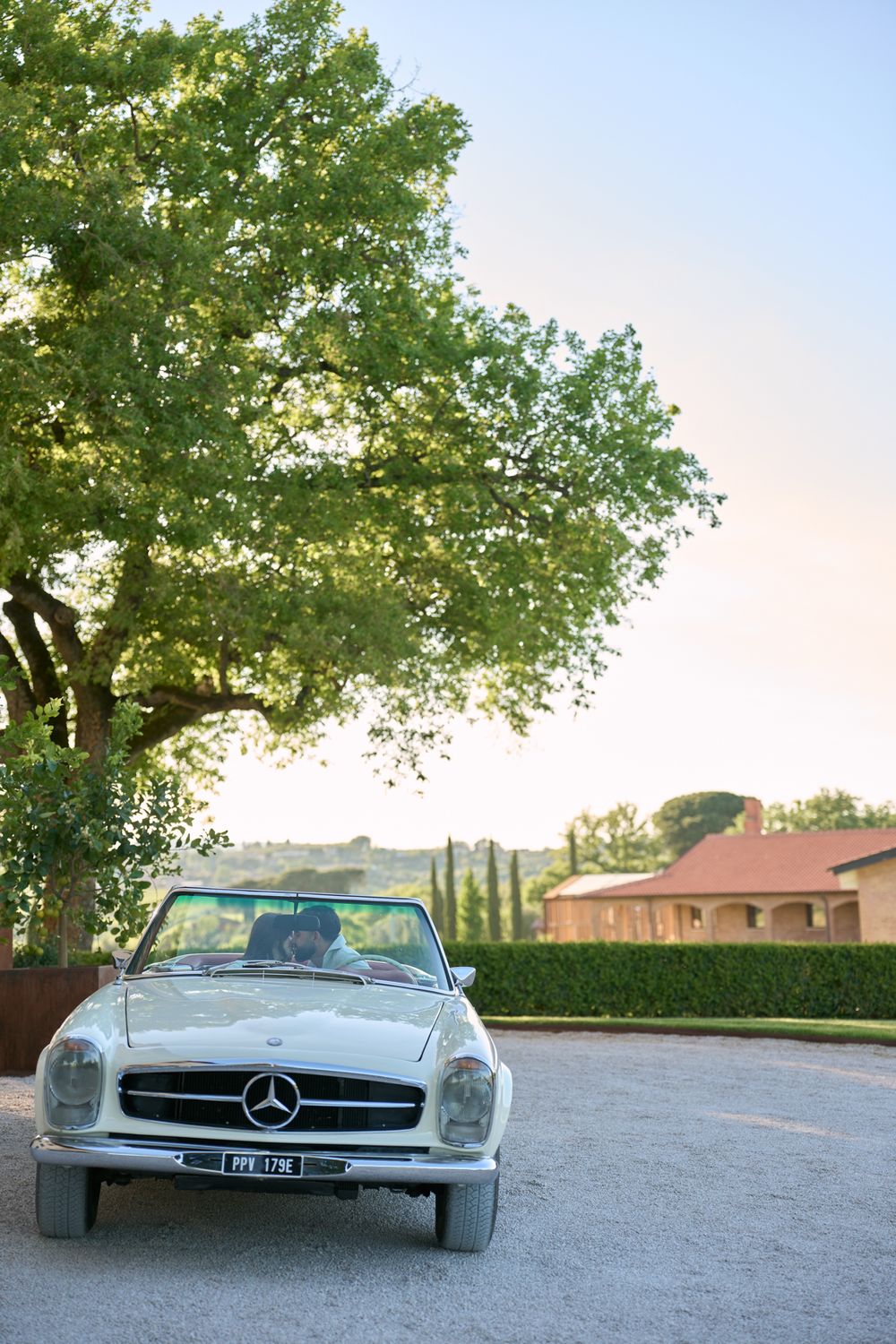 Classic white Mercedes-Benz convertible parked on gravel driveway surrounded by lush trees and Tuscan countryside landscape.