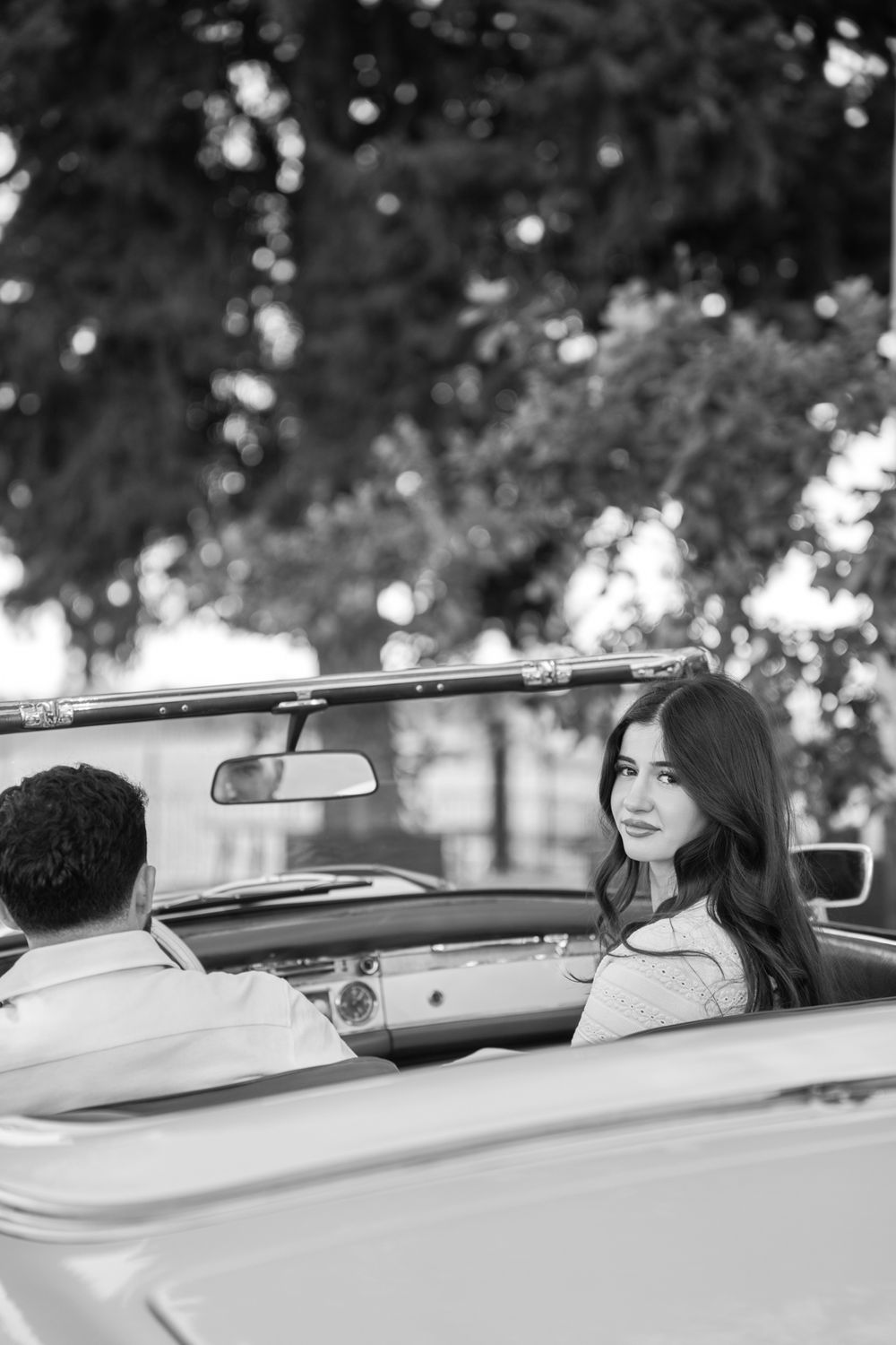 Black and white photo of a young woman with long hair sitting in a vintage convertible car outdoors.