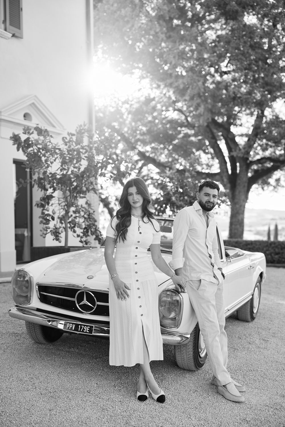 Black and white photo of a couple posing by a vintage Mercedes-Benz convertible with sunlight filtering through trees.