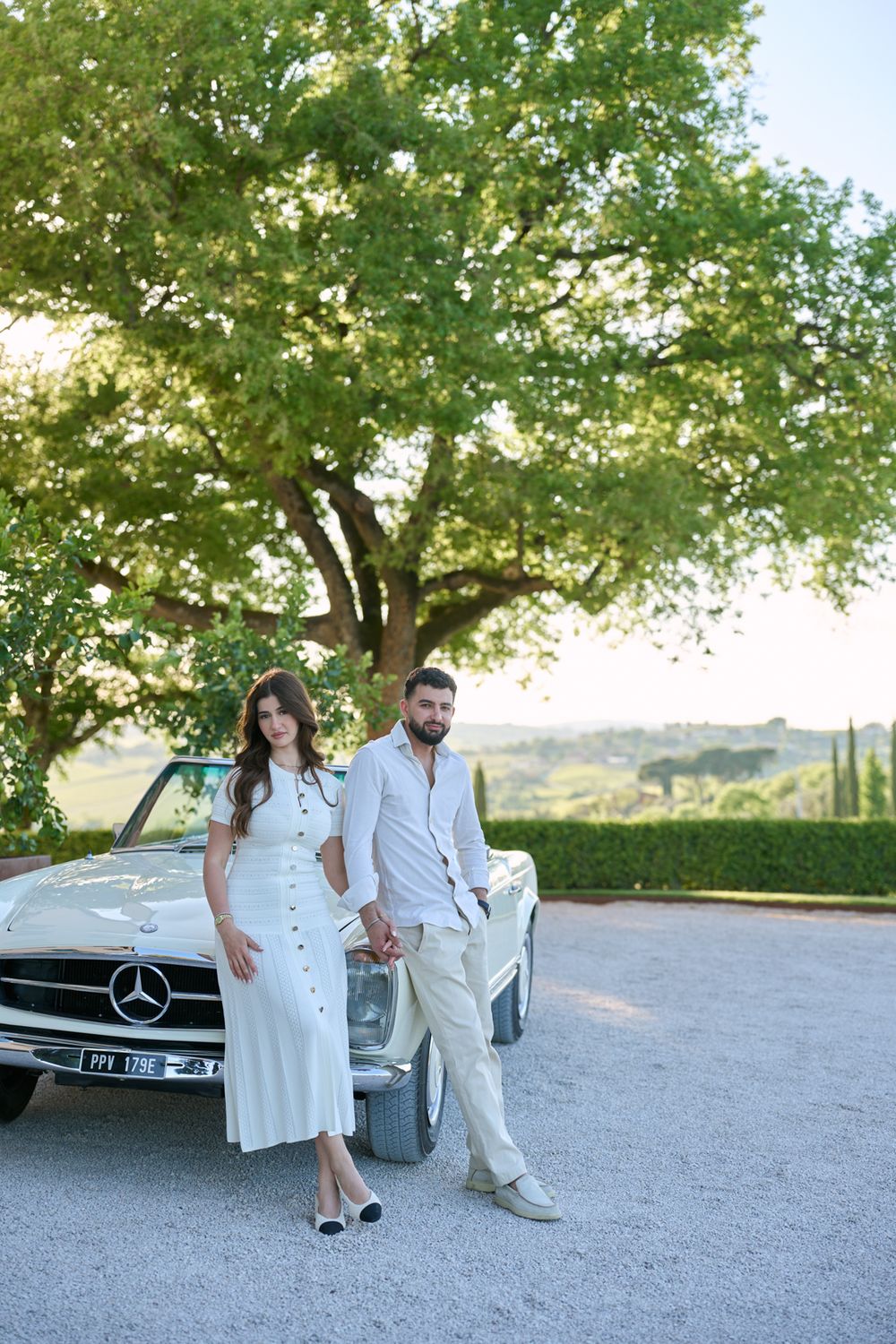 Elegant couple in white outfits posing by a classic Mercedes convertible under a large tree in Tuscany.