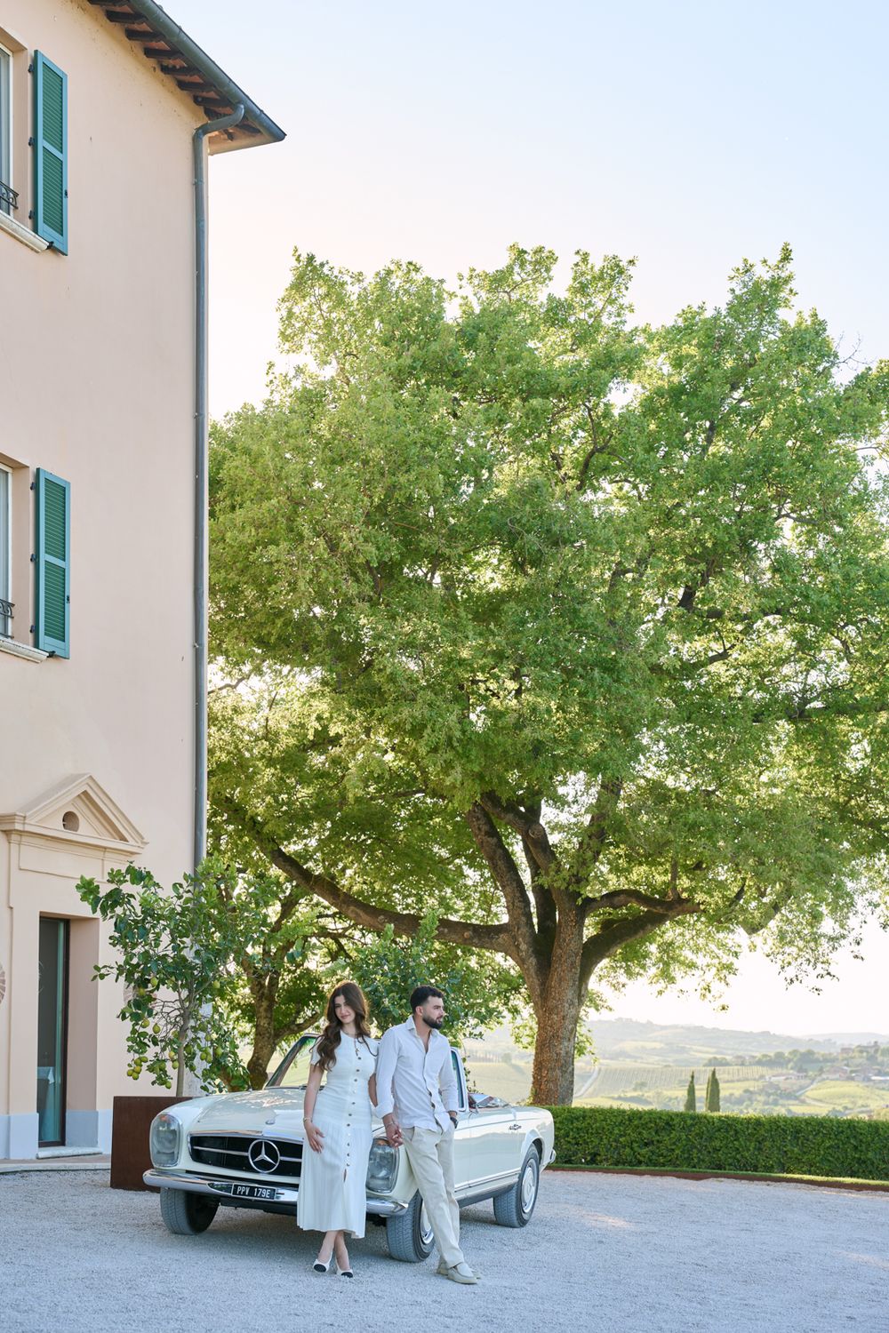 Couple in white attire leaning on a vintage Mercedes convertible outside a Tuscan villa with a large green tree.