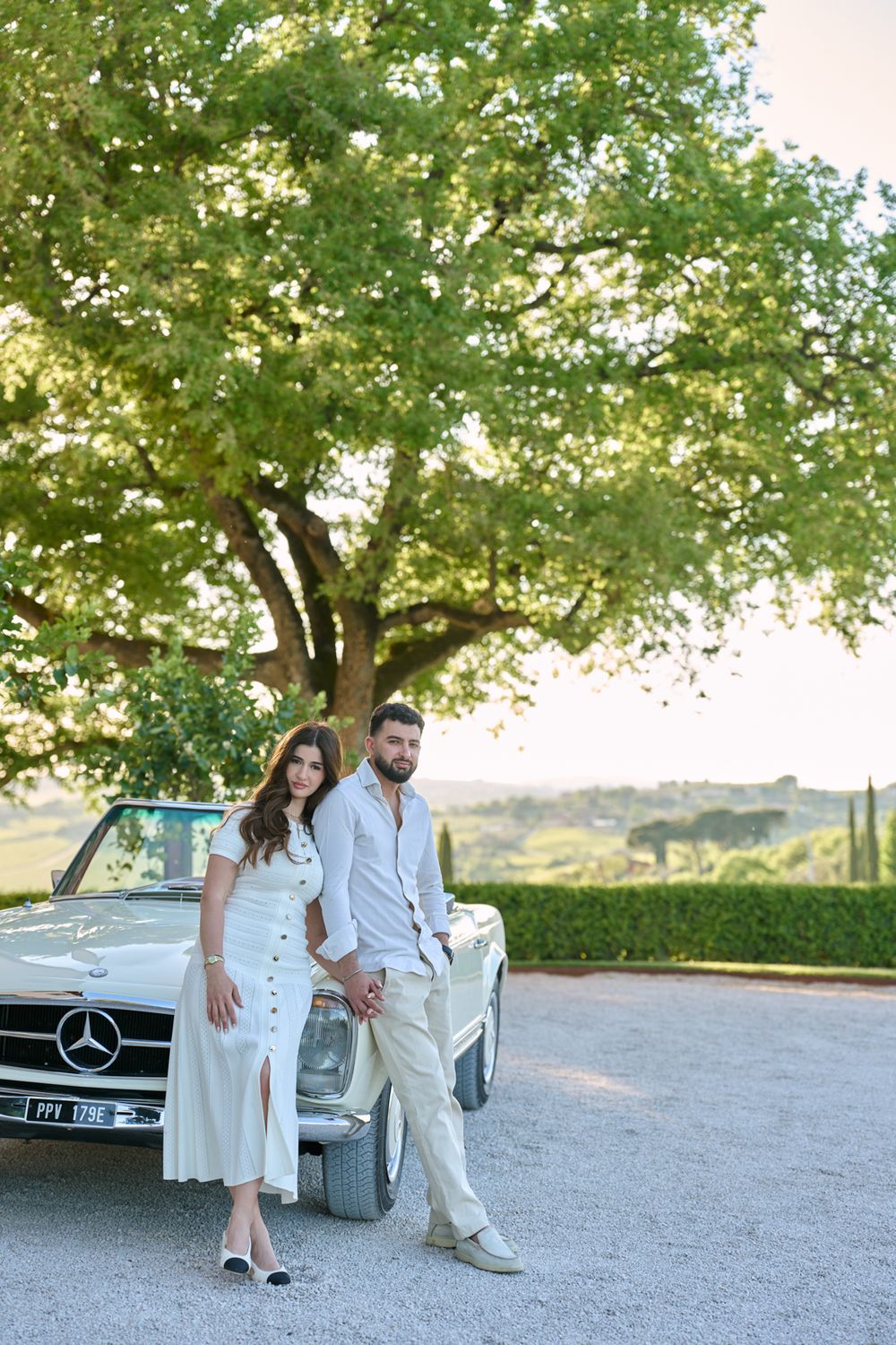 Elegant couple in white outfits leaning against a classic Mercedes convertible with a scenic Tuscan landscape backdrop.