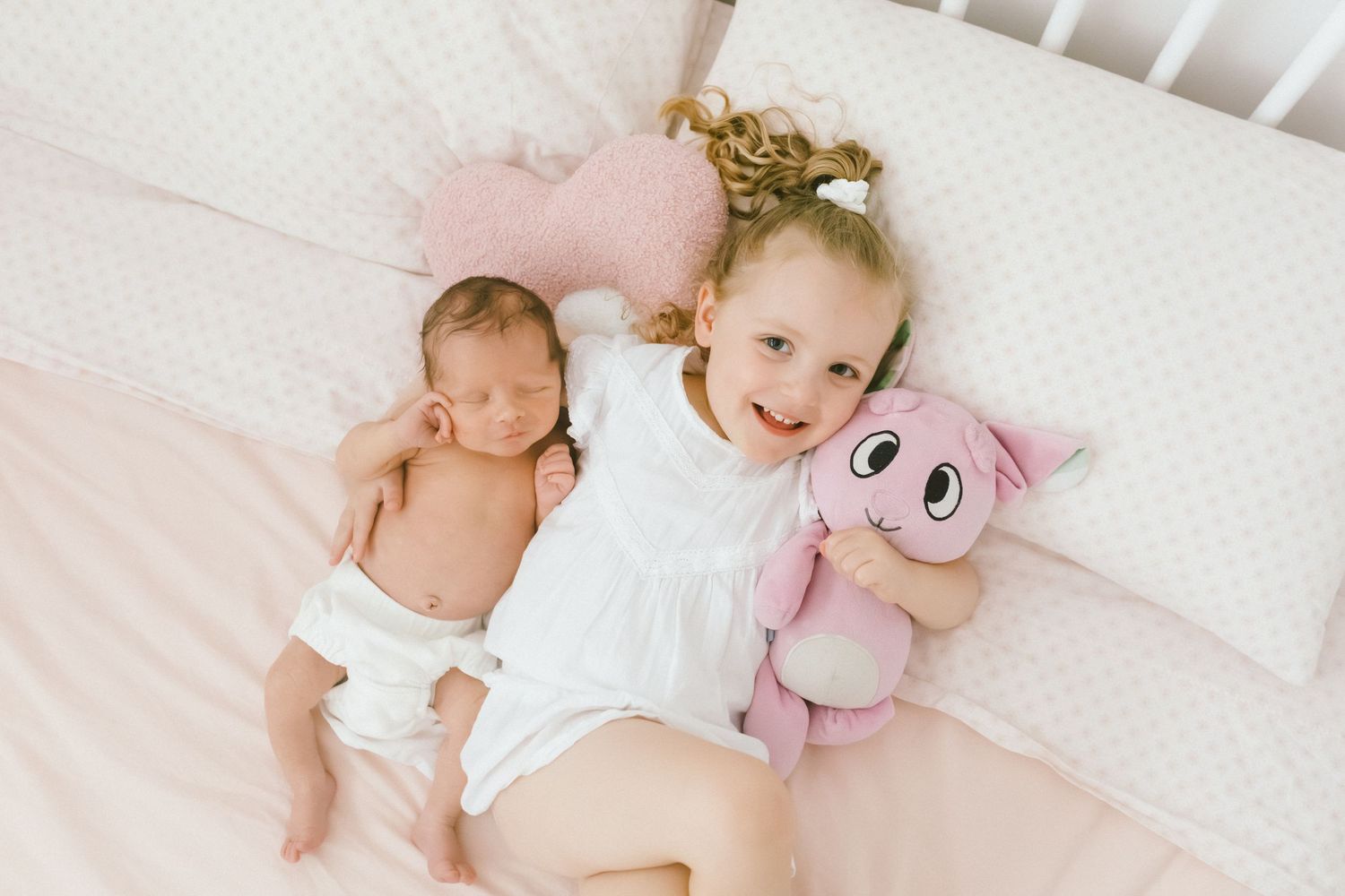 Young girl in white dress smiles holding pink stuffed animal beside sleeping newborn baby in a pink crib.