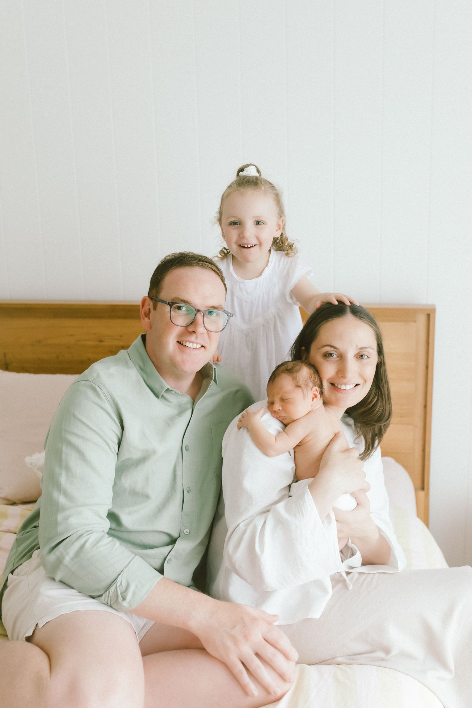 Happy family of four sitting on a bed, parents smiling with a toddler girl and newborn baby in a bright room.
