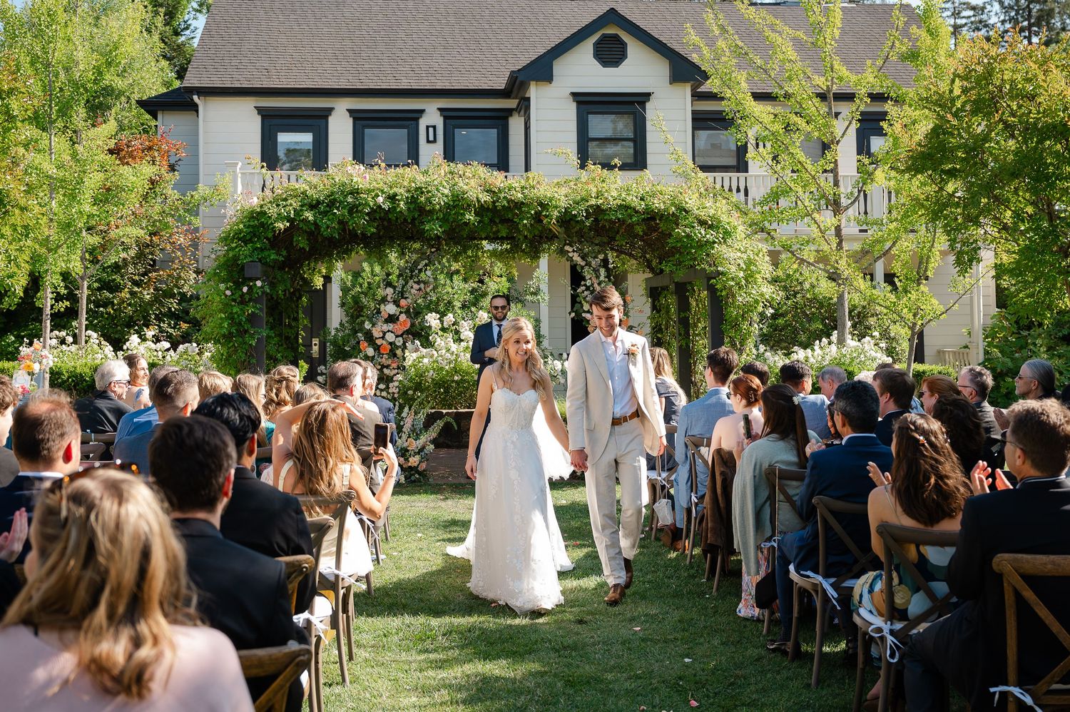 Guests dining at long outdoor tables on green lawn during elegant garden party wedding reception.