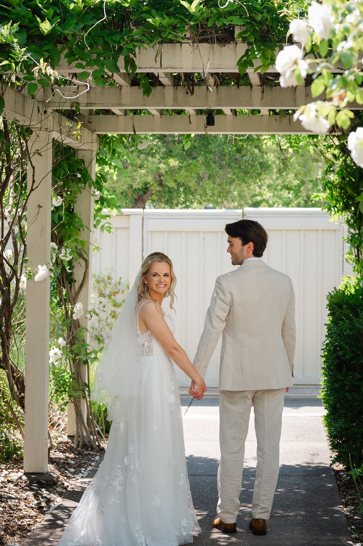 Bride in floral gown holds groom's hand under vine-covered pergola in lush garden wedding portrait.