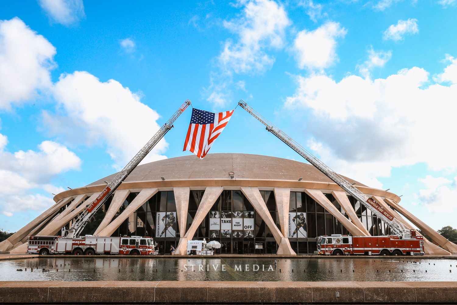 American flag flies between two fire truck ladders in front of a large domed building outdoors.