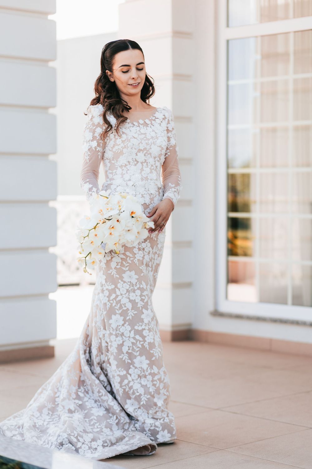 Beautiful bride in long-sleeve lace wedding gown holding white orchid bouquet outdoors.