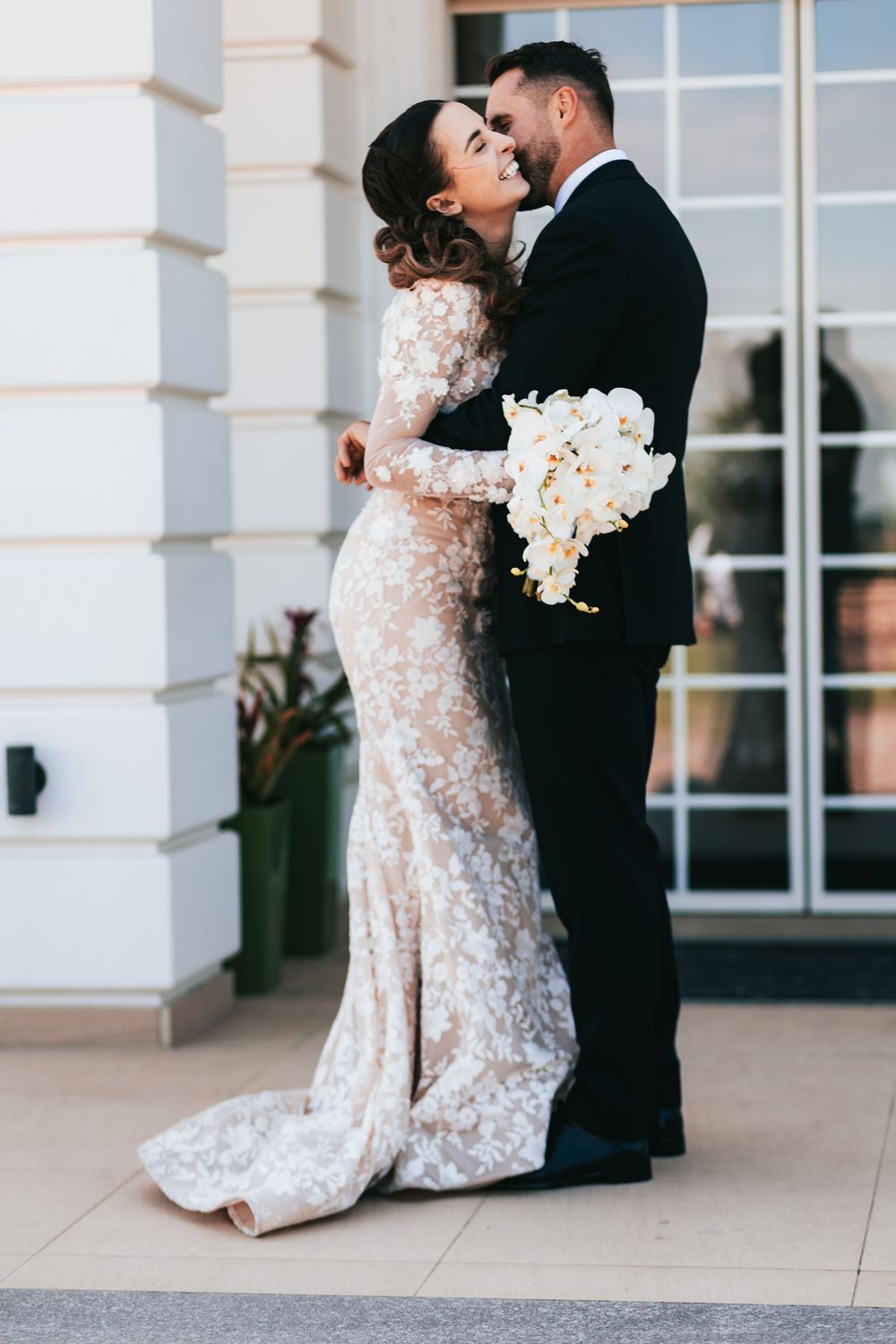 Bride in lace long-sleeve wedding gown embraces groom in black tuxedo outdoors holding white floral bouquet.