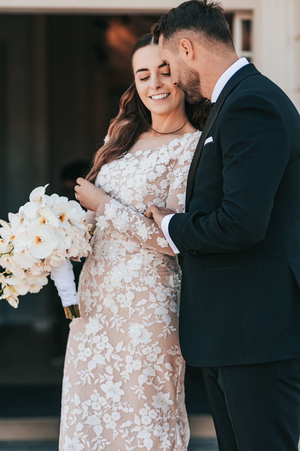 Smiling bride in floral lace gown holds white bouquet as groom in tuxedo whispers in her ear.