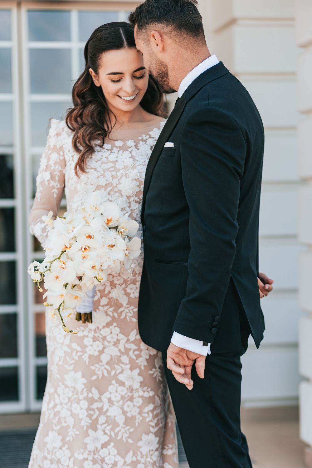 Groom in black tuxedo tenderly kissing bride wearing lace wedding dress while she holds white orchid bouquet.