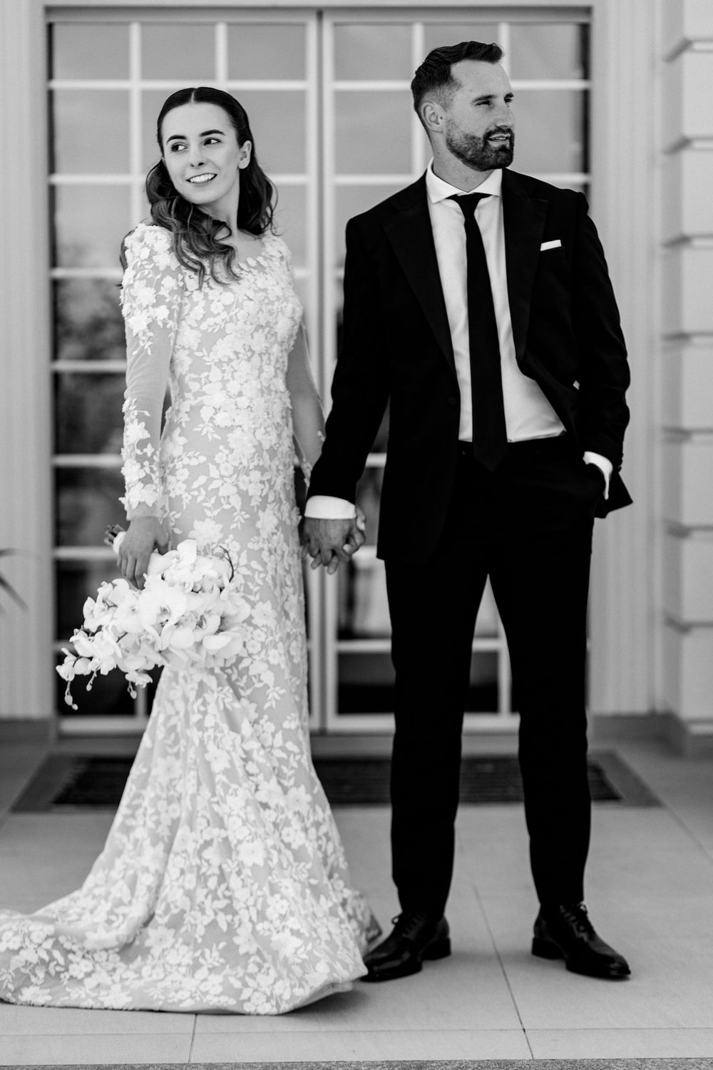 Black and white portrait of bride in lace mermaid wedding dress and groom in black suit holding hands outdoors.