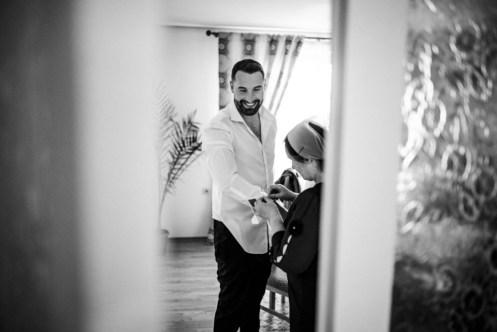 Black and white photo of groom in white shirt being helped with jacket by wedding assistant indoors.
