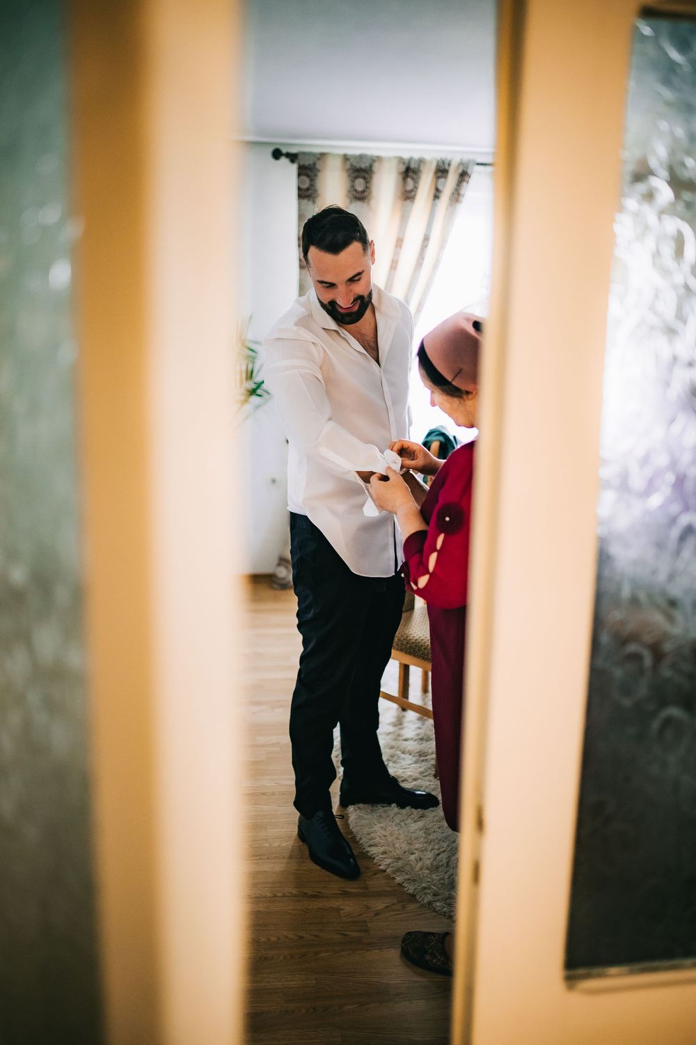 Groom in white shirt getting ready with help from family member in cozy home interior before wedding.