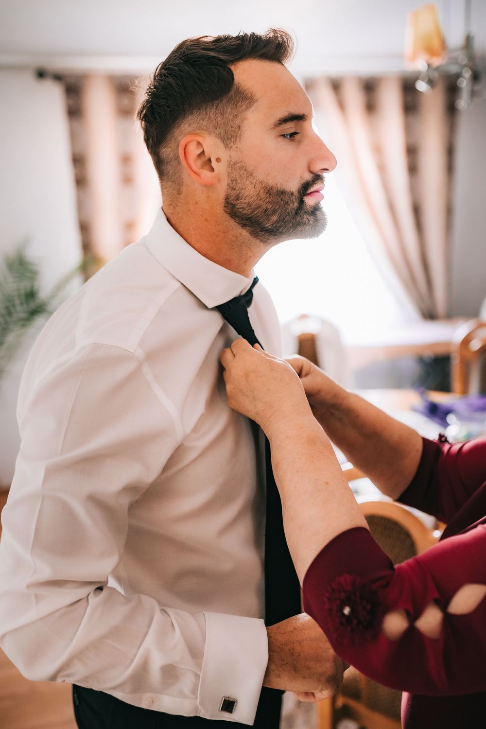 Groom in white dress shirt having his black tie adjusted by someone during wedding day preparations indoors.