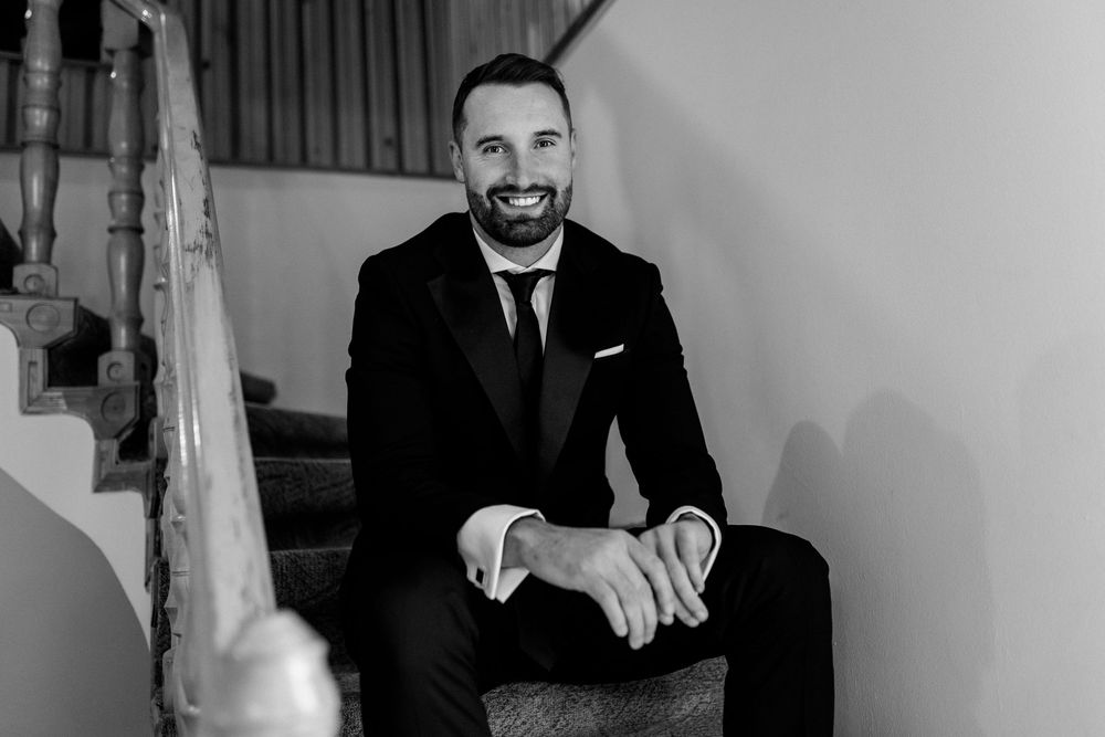 Handsome groom in black tuxedo smiling while sitting on staircase in black and white wedding photo.