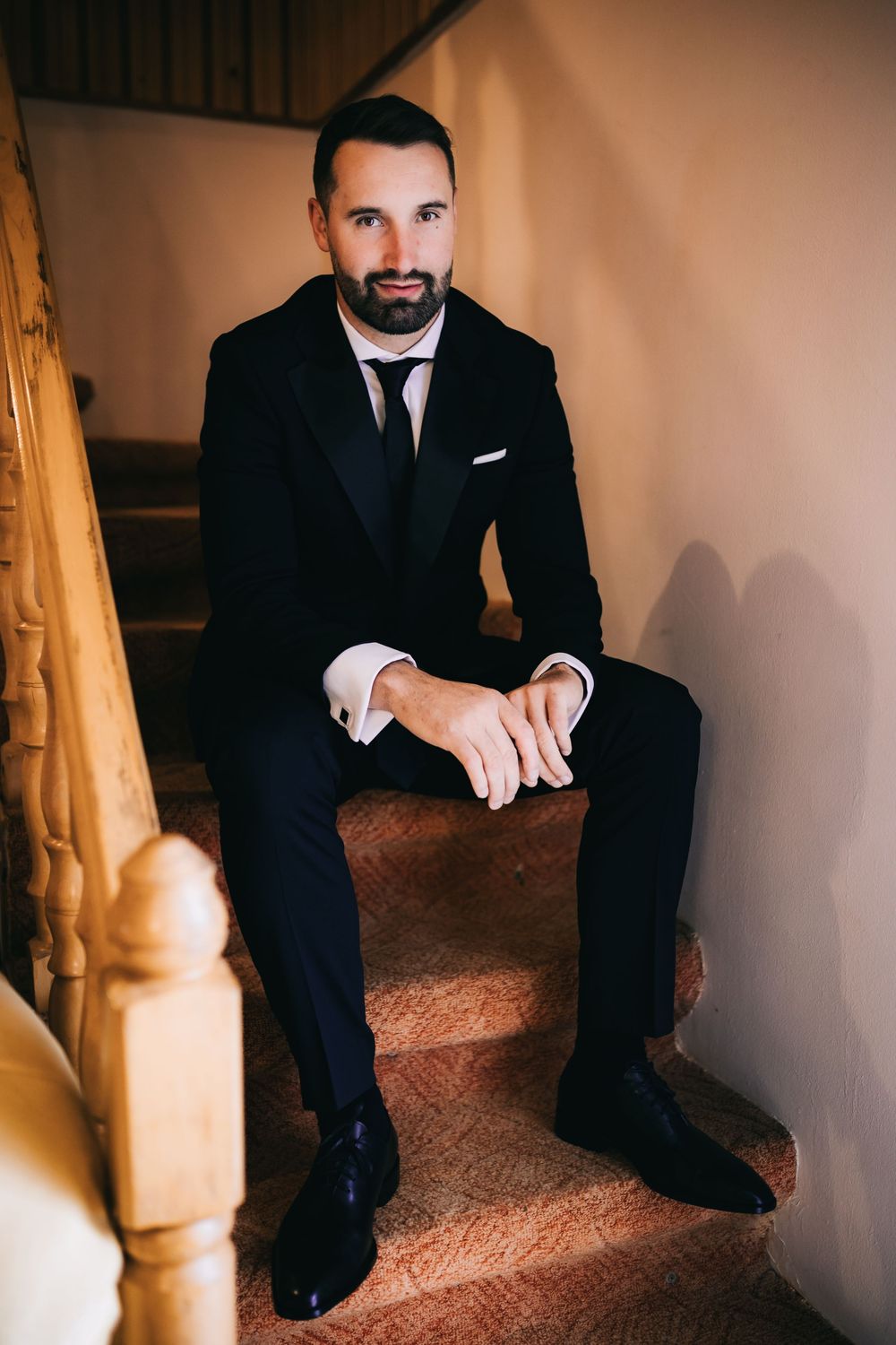 Groom in black tuxedo sitting on carpeted staircase, hands clasped, looking confident before wedding ceremony.
